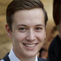 Young man with short brown hair smiling, wearing a suit jacket and white shirt, blurred warm-toned background.