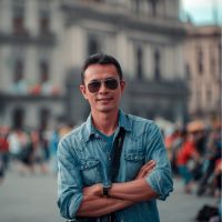 Man wearing sunglasses and a denim shirt with arms crossed, standing in a busy city square with blurred historic buildings.