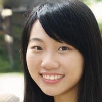 Young woman with long dark hair smiling at the camera against a softly blurred outdoor background.