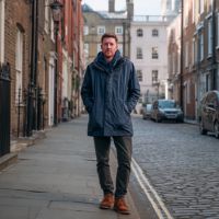 Man in navy coat with hands in pockets walking on a cobblestone street lined with brick buildings and parked cars.