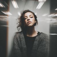 Woman with wavy hair wearing a gray cardigan, gazing at the camera amid radial light streaks and overhead fluorescent lights.