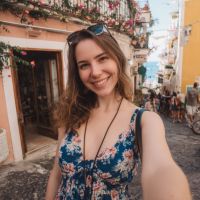 Smiling woman in a floral dress taking a selfie on a sunlit street with colorful buildings and flowers.