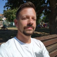 Man with short hair and goatee sitting on a park bench, wearing a white t-shirt and squinting in sunlight.
