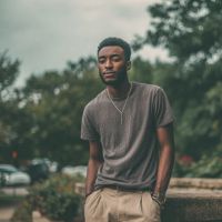 Young man outdoors with hands in pockets, wearing a gray t-shirt, beige pants and pendant necklace against blurred trees.