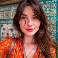 Young woman with long brown hair in an orange patterned blouse, smiling in front of an ornate blue-and-yellow tiled wall