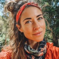 Woman with red headband and messy bun wearing a floral neck gaiter and orange jacket, smiling outdoors with trees behind.