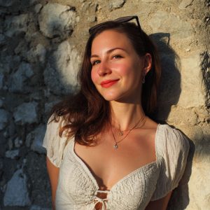Young woman with sunglasses on her head, smiling against a sunlit stone wall, wearing a white lace top and necklace.