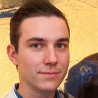 Man with short brown hair and neutral expression wearing a collared shirt against a warm yellow background