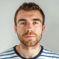 Adult man with short brown curly hair and light stubble, wearing a white shirt with blue stripes and a neutral expression.