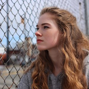 Young woman with long wavy red hair and freckles gazes past a chain-link fence toward a blurred urban background.