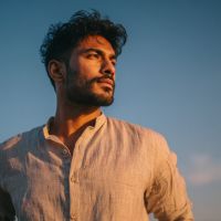 Bearded man with dark curly hair in a light linen shirt gazing into sunlight against a clear blue sky.
