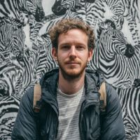 Young bearded man with curly hair wearing a jacket and backpack, standing against a black-and-white zebra mural.