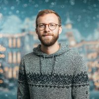 Young man with glasses and short beard wearing a gray patterned knit hoodie against a blurred festive bokeh background.