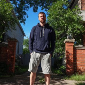 Bearded man in a navy hoodie and light gray shorts stands with hands in pockets between brick pillars on a sunny sidewalk.