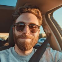 Bearded man wearing sunglasses and a seatbelt smiling inside a sunlit car.