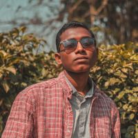 Young man with short hair and sunglasses wearing a red plaid shirt over a gray button-down, standing before leafy shrubs.
