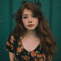 Person with long wavy red hair wearing a black floral dress, looking at the camera against a teal wooden door.