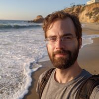 Bearded man with glasses and a backpack smiling at the camera on a sandy beach with waves and coastal cliffs.