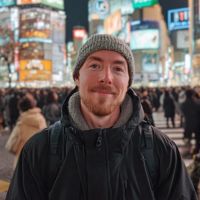 Man with a gray knit beanie and reddish beard smiling in a crowded, neon-lit city street at night.