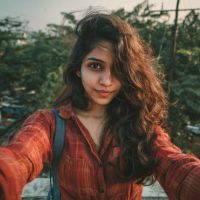 Young woman with long wavy brown hair in a red striped shirt and backpack strap, taking a selfie outdoors with trees behind her.