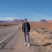 Person wearing a dark jacket, gray pants and white sneakers standing on a deserted road with red-rock mesas under a clear blue sky.