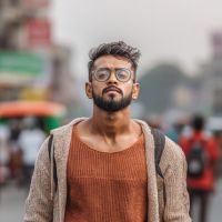 Young man with glasses and beard wearing a rust sweater and backpack standing on a busy street.