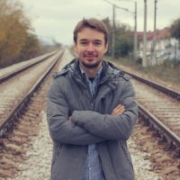 Man standing on railroad tracks with arms crossed, wearing a gray jacket and blue shirt, slight smile, houses and trees in the background.